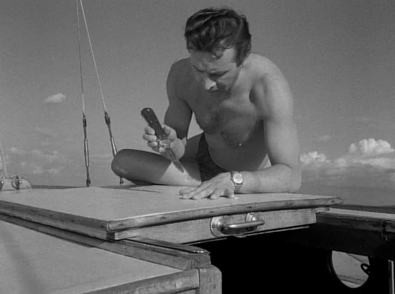 Knife In The Water (1962) movie still: medium — A wooden hatch cover on the deck of a sailboat under a clear sky. A shirtless man sits on the deck…