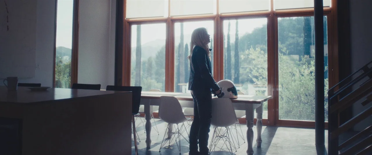 Gemini (2017) movie still: wide — A spacious room features floor-to-ceiling windows with wooden frames and white Eames-style chairs. A blonde woman…