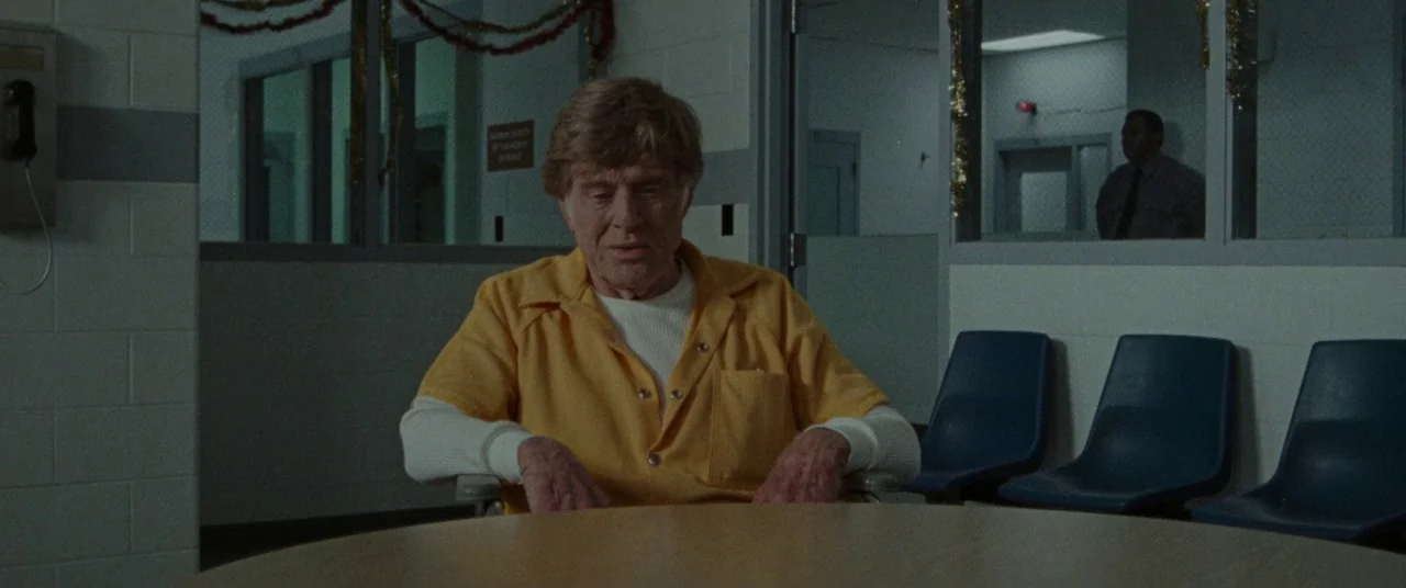 The Old Man & The Gun (2018) movie still: medium — A austere room with cinder block walls, plastic blue chairs, and glass partitions. An elderly man…