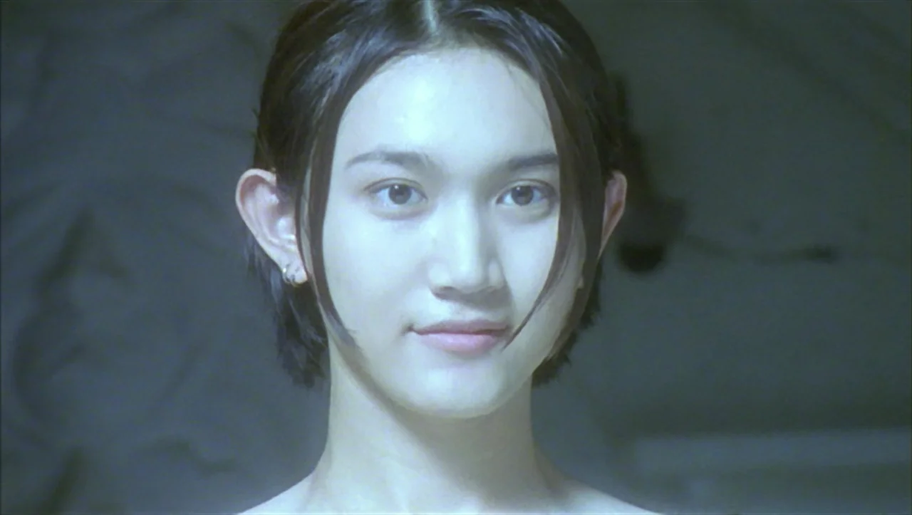 Wild Zero (1999) movie still: closeup — A person is centered in a brightly lit, undifferentiated interior space with neutral tones. The subject…