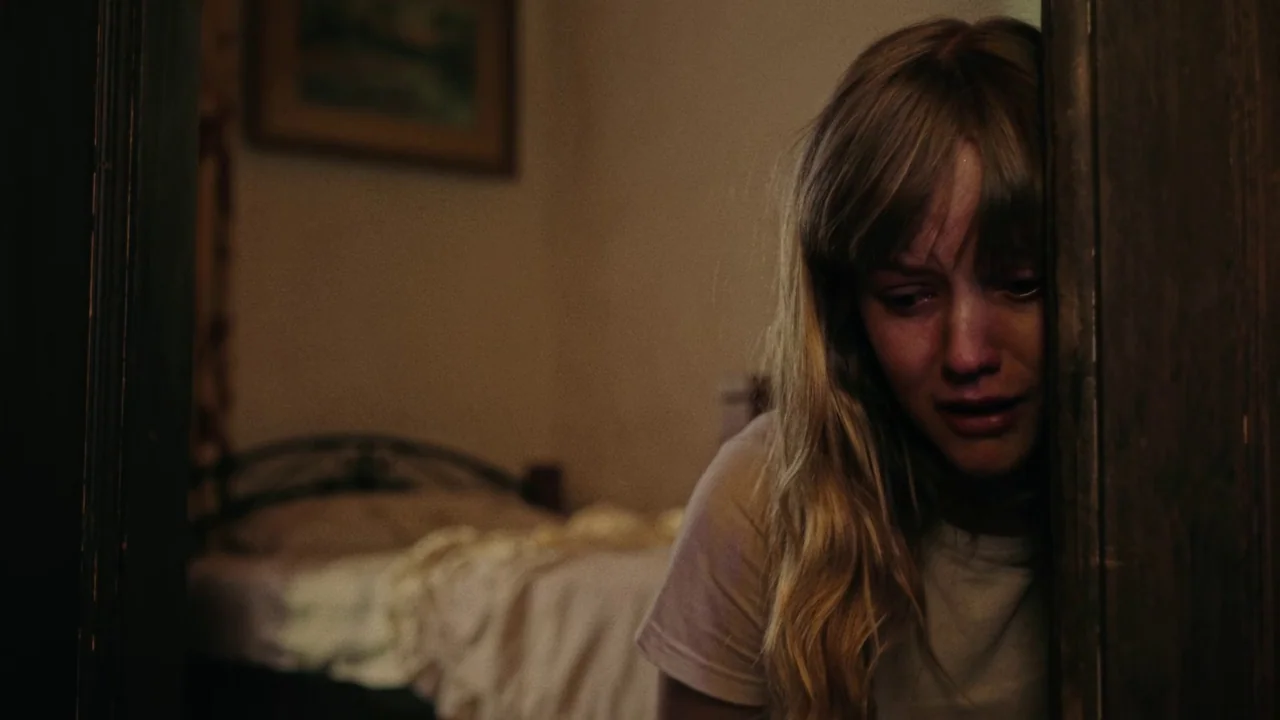 My First Summer (2020) movie still: closeup — A dimly lit room with an unmade bed visible in the blurred background. A young woman stands partially…