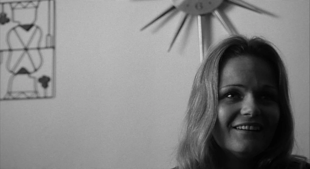 Lenny (1974) movie still: closeup — A room with white walls featuring an abstract metal wall sculpture and starburst clock. A young woman turns her…