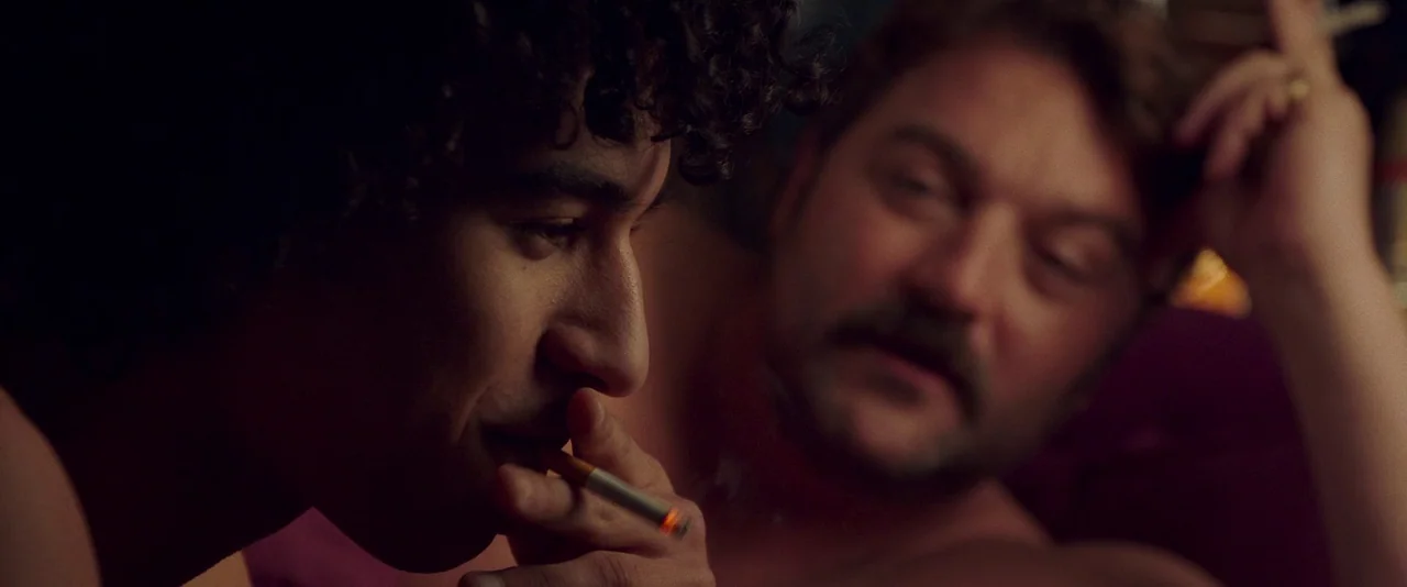 Peter Von Kant (2022) movie still: closeup — A dimly lit room with a deep red background texture. Two men are lying in bed together while one holds…