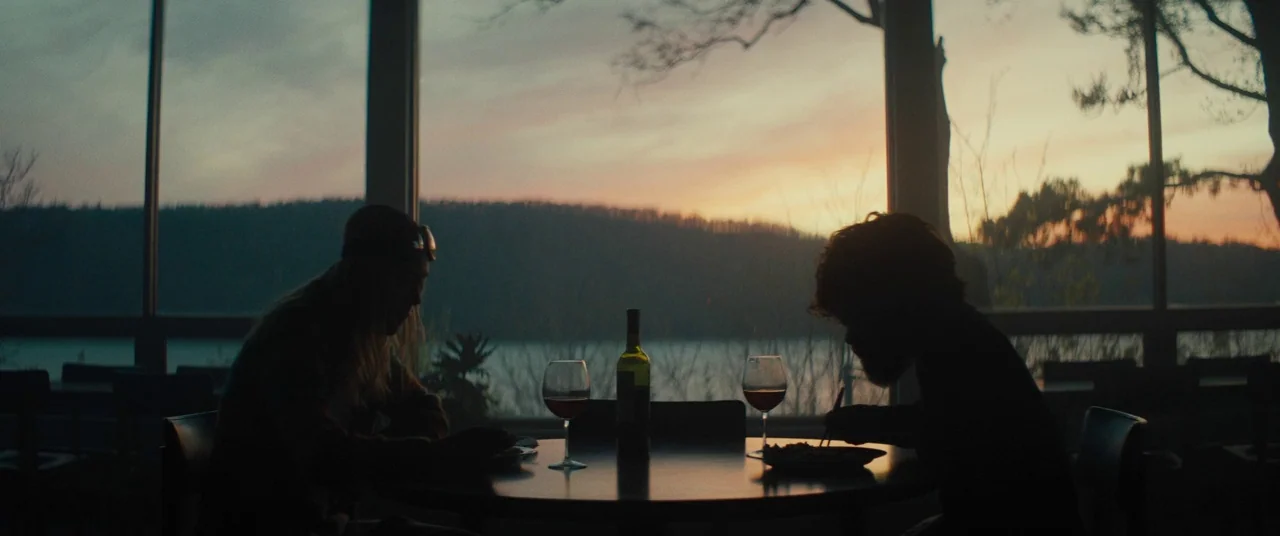 I Think We’re Alone Now (2018) movie still: wide — Two individuals sit at a circular table in front of a floor-to-ceiling window. They eat dinner…