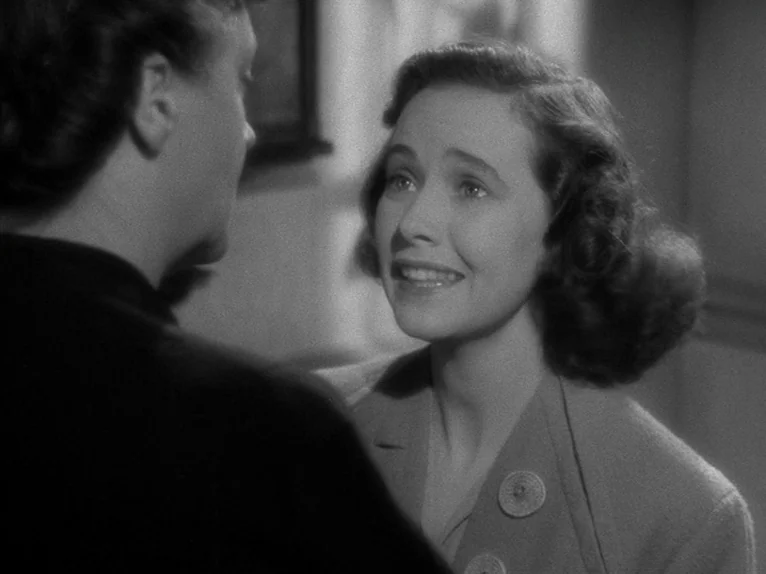 Shadow of a Doubt (1943) movie still: medium — A woman stands in a bright room and speaks to a man seen from behind. The man in a dark coat faces…