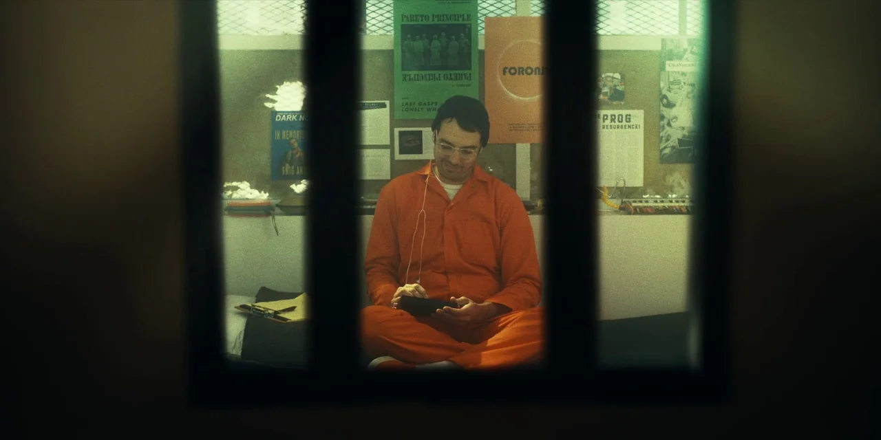 Destroy All Neighbours (2024) movie still: medium — A spartan room features beige walls covered in various posters and pinned documents. A man in an…