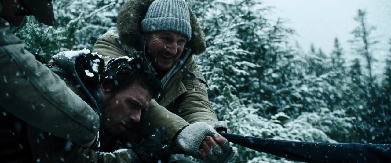 The Grey (2012) movie still: medium — A dense forest landscape with heavy snowfall covering pine branches during a winter day. Two men grip a black…