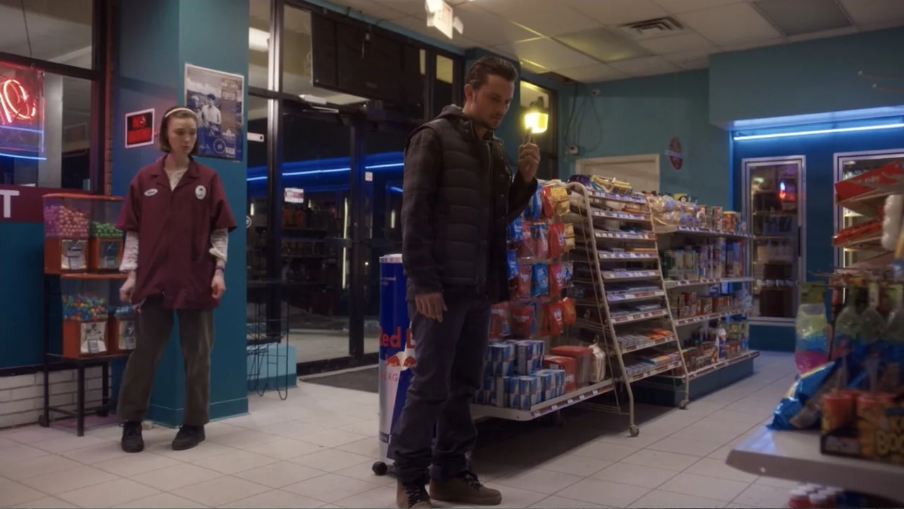 Burn (2019) movie still: fullbody — A brightly lit convenience store interior features blue walls, tiled floors, and product shelves. A young man…
