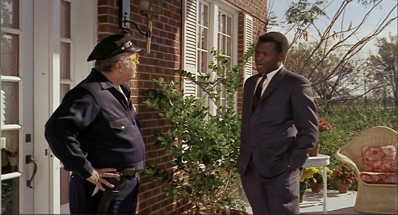In the Heat of the Night (1967) movie still: medium — A red brick residential exterior features a white-framed doorway and wicker furniture. Two men…