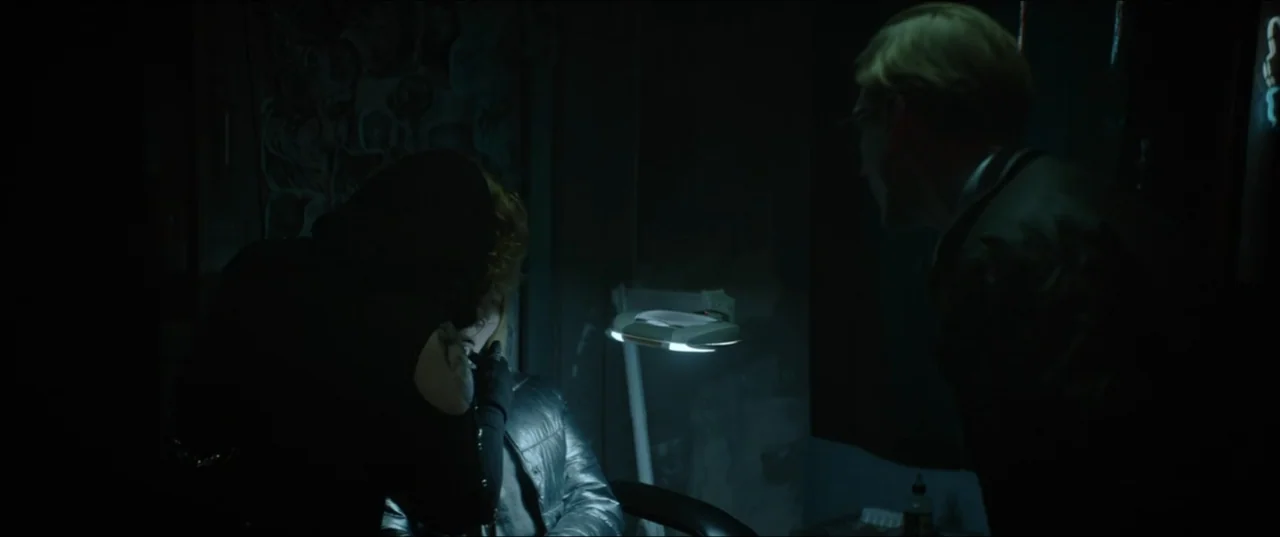 Dogs Don't Wear Pants (2019) movie still: medium — A dark, dimly lit professional space with a prominent adjustable magnifying lamp overhead. Two…
