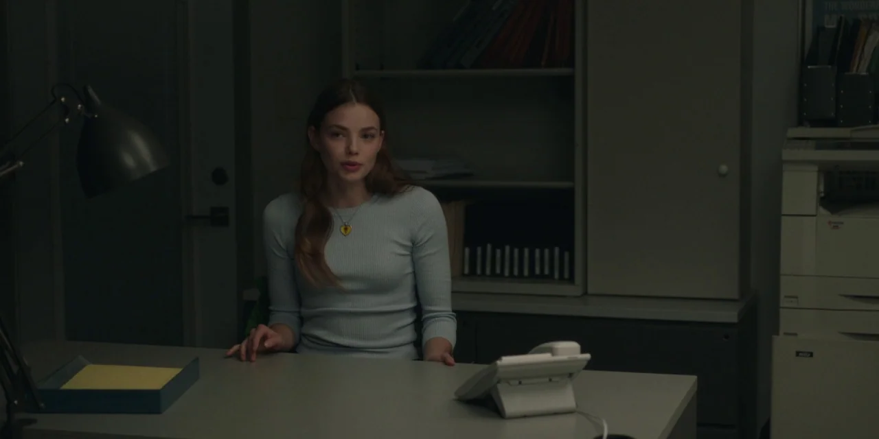 The Assistant (2019) movie still: medium — A dimly lit room containing a desk, a copier, and storage shelving. A woman sits at the desk looking…