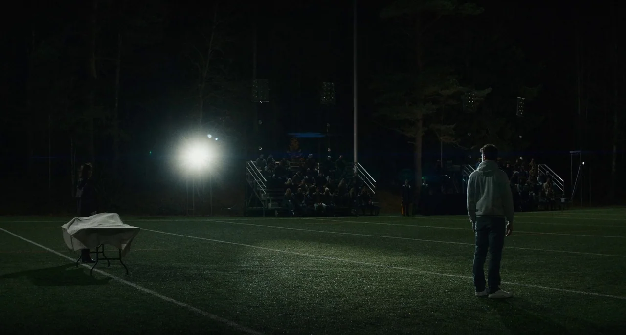 Dual (2022) movie still: wide — A night scene on an empty sports field illuminated by bright stadium lights. A male student stands on the turf…