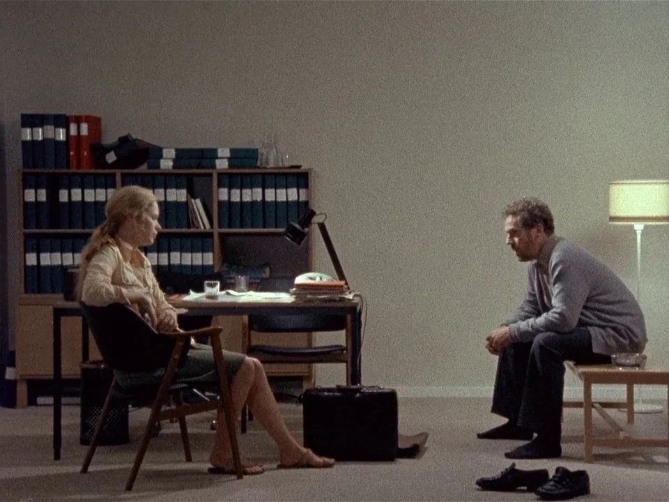 Scenes from a Marriage (1973) movie still: wide — A spartan room features a bookshelf with many blue folders and sparse furniture. A woman sits in a…