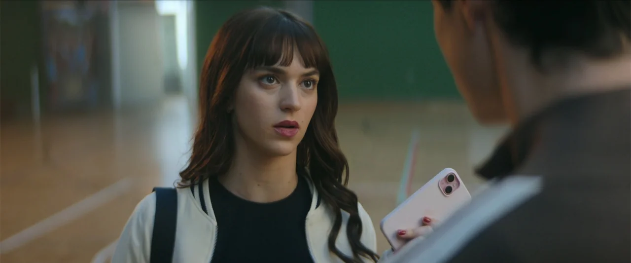 Tell Me Softly (2025) movie still: medium — An interior sports hall features a green wall and a hardwood floor. A young woman with bangs holds a…