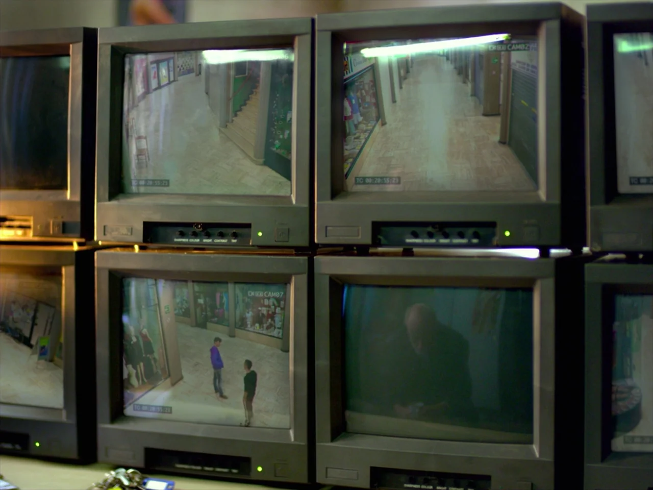 Violet (2014) movie still: medium — A wall of stacked cathode ray tube monitors displays surveillance footage from a mall. Each screen shows…
