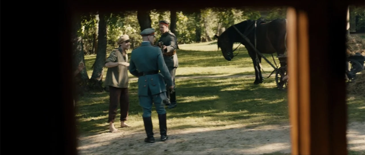 The Rifleman (2019) movie still: medium — A rural setting with green grass and trees during a sunny day. Two soldiers in uniforms stand and talk…