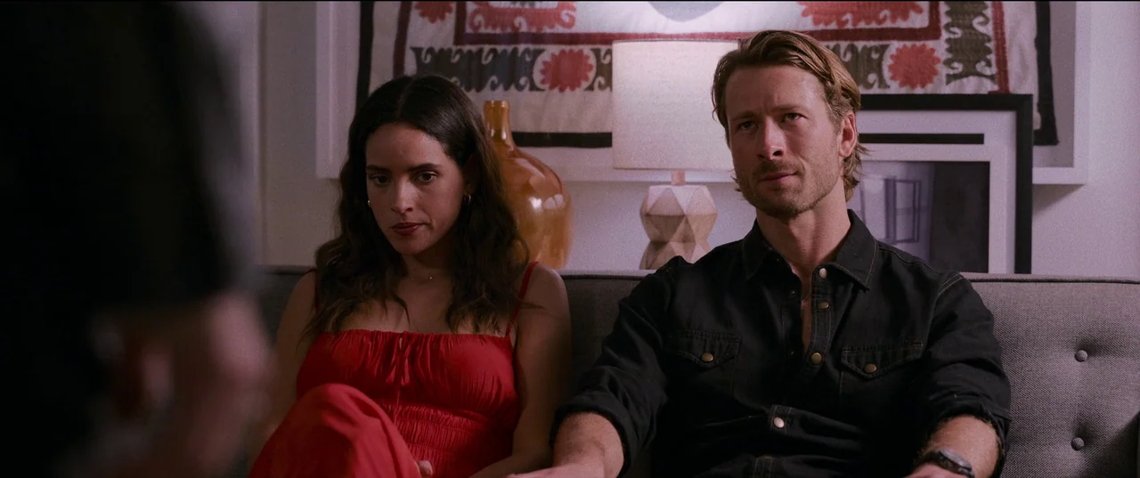 Hit Man (2023) movie still: medium — A man and woman sit side by side on a neutral-toned sofa. A decorative wall hanging and a table lamp sit behind…