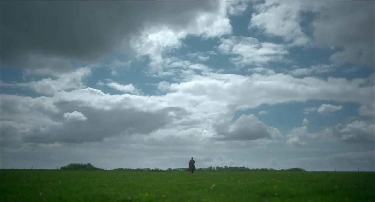 Men (2022) movie still: wide — A vast, open green meadow stretches out under a sprawling, cloudy daytime sky. A solitary figure wearing a long coat…