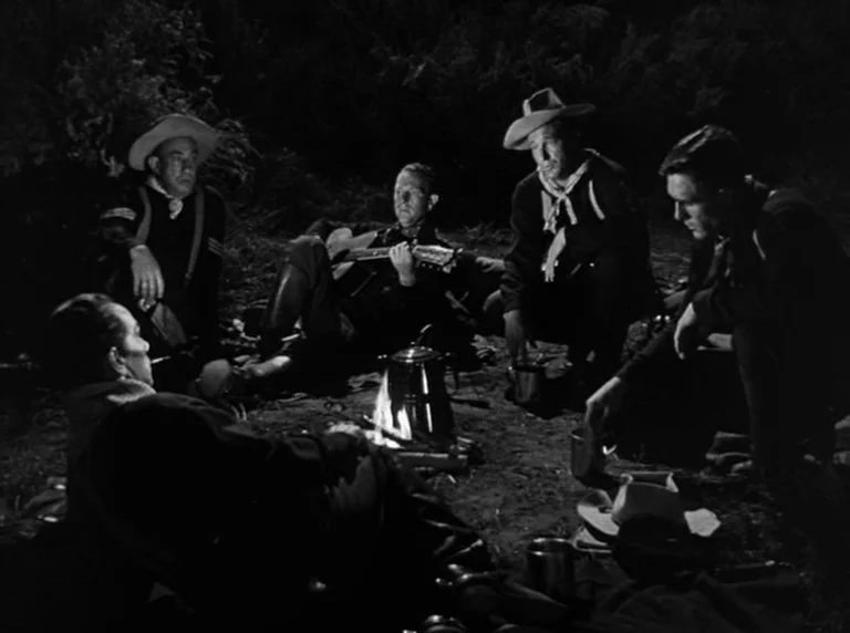 Rio Grande (1950) movie still: medium — Four men sit on the ground at night surrounding a small, glowing campfire. One man plays an acoustic guitar…