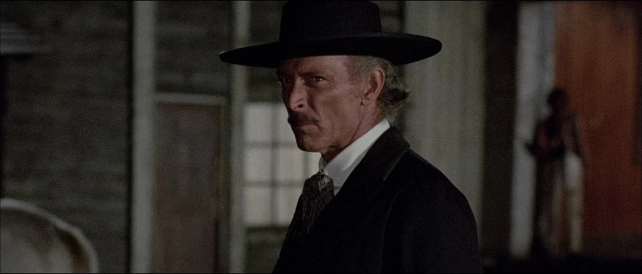 The Grand Duel (1972) movie still: medium — A dimly lit room with simple wood-paneled walls and a soft glow in the background. A man in a western…