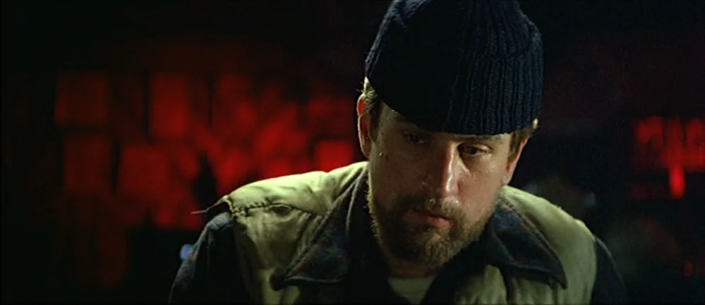 The Deer Hunter (1978) movie still: closeup — A dimly lit interior space with red neon light cast across the background. A man wearing a navy beanie…