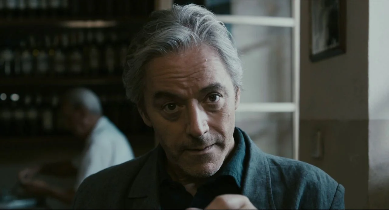 Certified Copy (2010) movie still: closeup — A man stands in an indoor cafe with shelves filled with bottles in background. The man looks forward at…