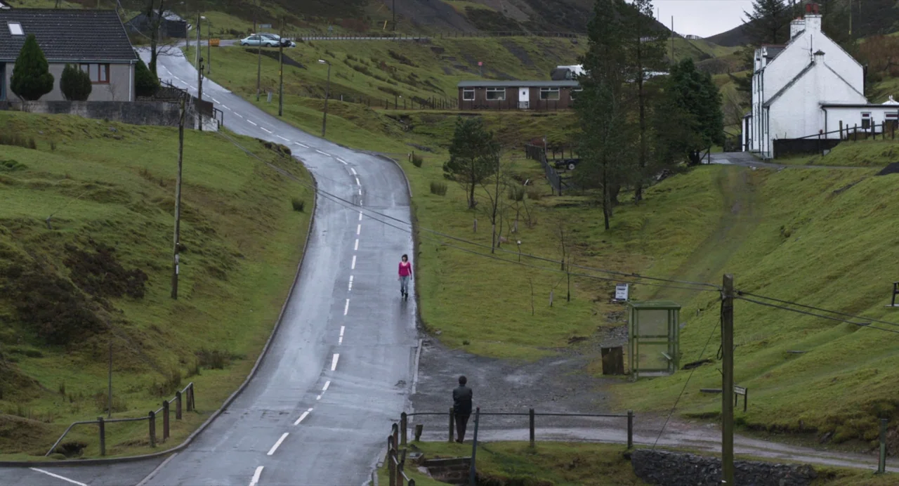 Under The Skin (2013) movie still: wide — A winding asphalt road cuts through a hilly, green rural landscape under overcast skies. One person walks…