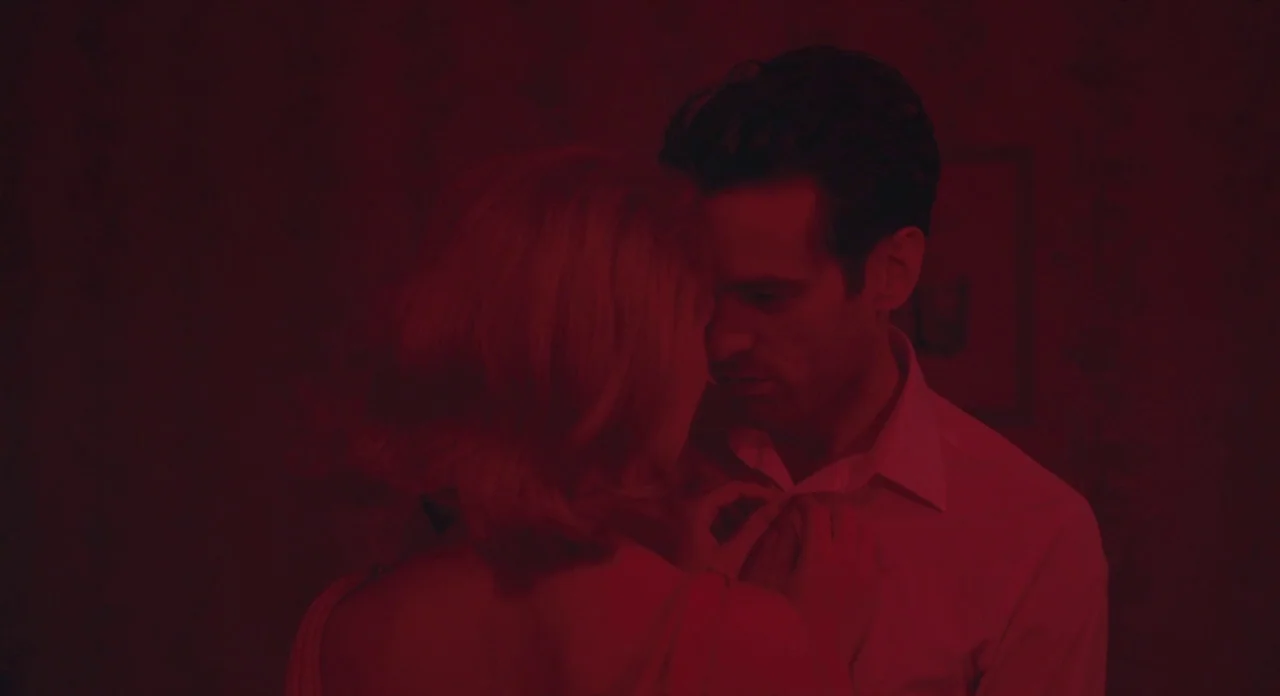 Populaire (2012) movie still: medium — A man and woman stand close together in a dark room illuminated by red light. The woman reaches up to adjust…