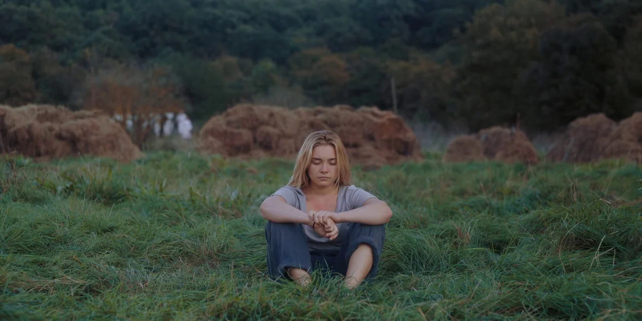 Midsommar (2019) movie still: medium — A woman sits in a field filled with tall grass and large hay bales. She sits with her legs tucked inward and…