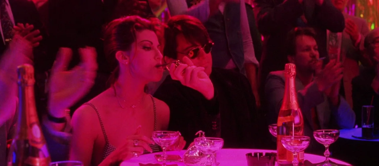Showgirls (1995) movie still: medium — A dimly lit nightclub features a table surrounded by people in dark attire. A woman and a man sit together at…