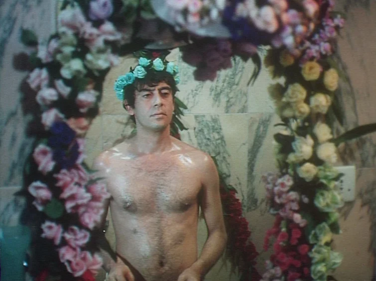 Hermitage (1968) movie still: medium — A bathroom vanity mirror is framed by decorative garlands of colorful artificial roses. A shirtless man…