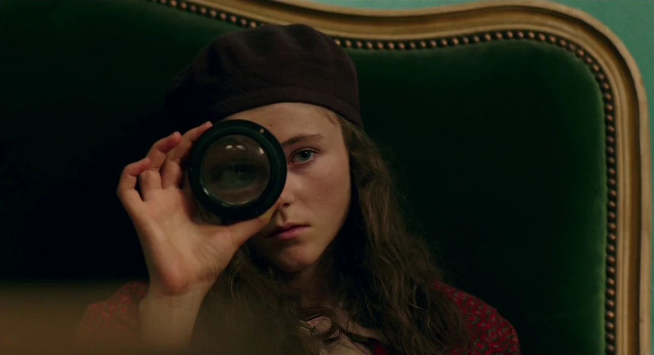 Jojo Rabbit (2019) movie still: closeup — A person sits in front of a green velvet chair with a decorative gold frame. The subject holds a circular…