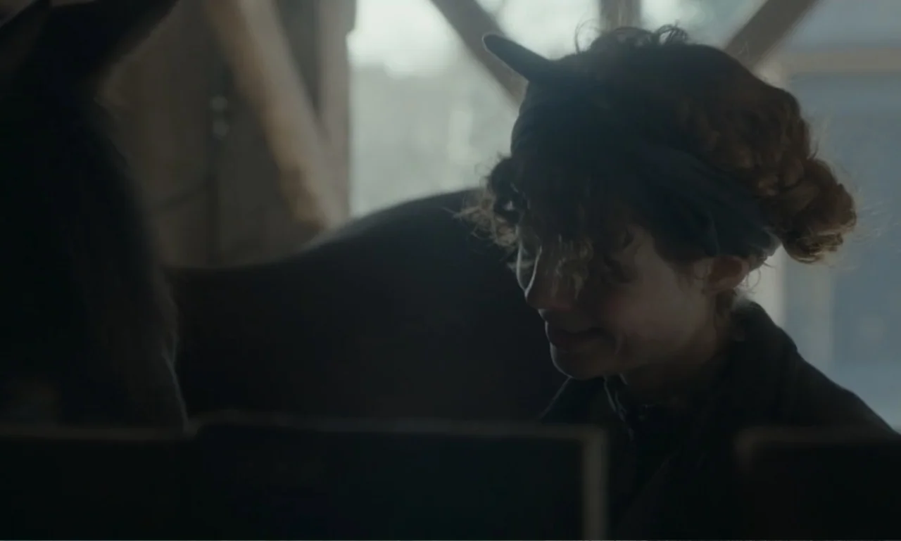 The Ugly Stepsister (2025) movie still: medium — A dim wooden stable interior with blurred structural beams in the background. A young woman stands…