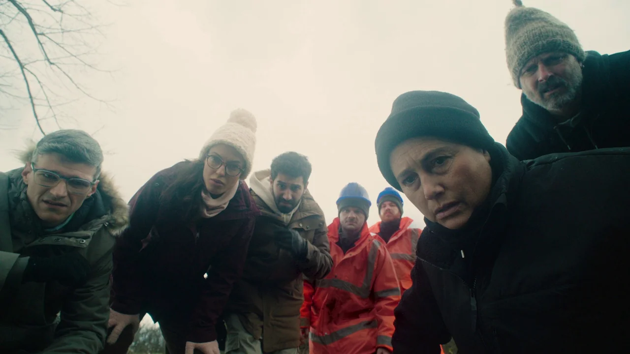 Helll Hole (2024) movie still: medium — A group of seven people cluster together in an outdoor area looking downward. They appear concerned as they…