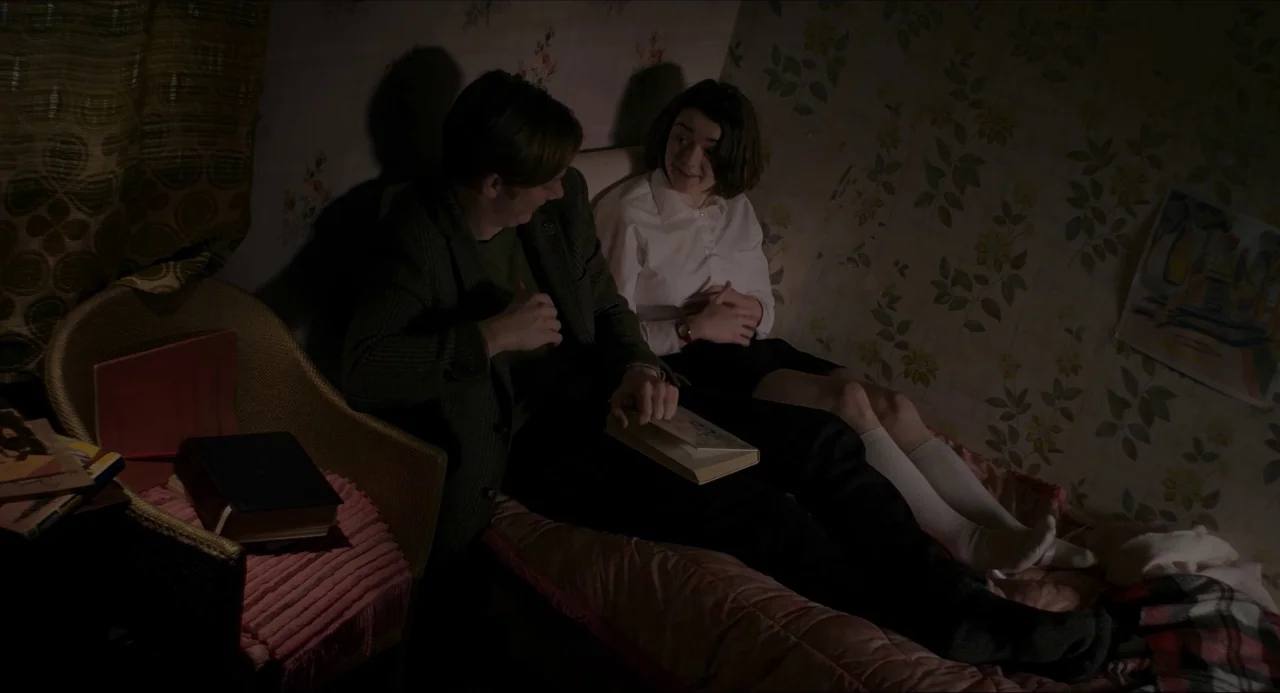 The Falling movie still: medium — A small room features floral wallpaper and a bed where a couple sits together. The man points to an open book…