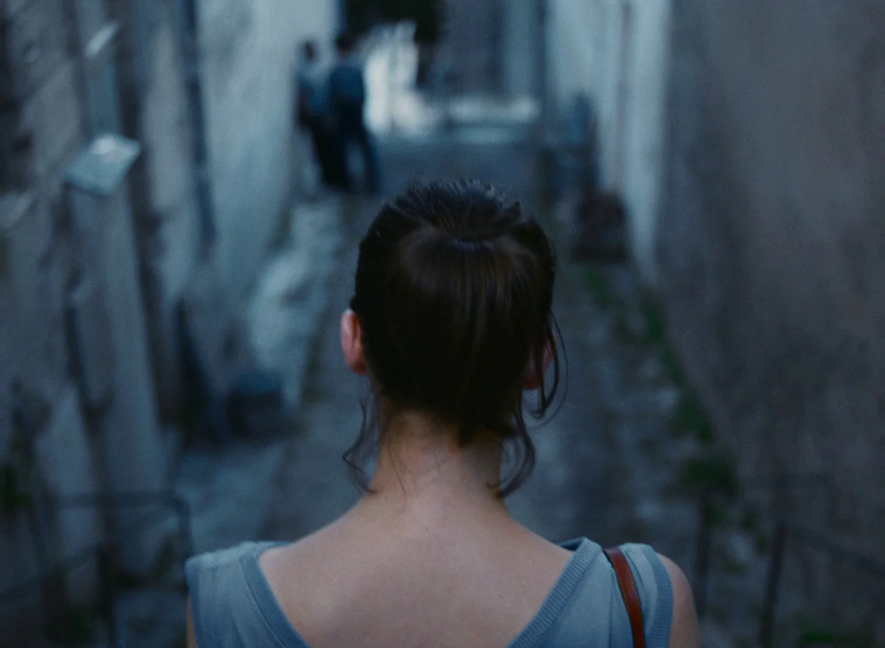 Happening (2021) movie still: medium — A narrow, stone-walled alleyway extends into the distance under cool, dim blue light. A woman walks away from…