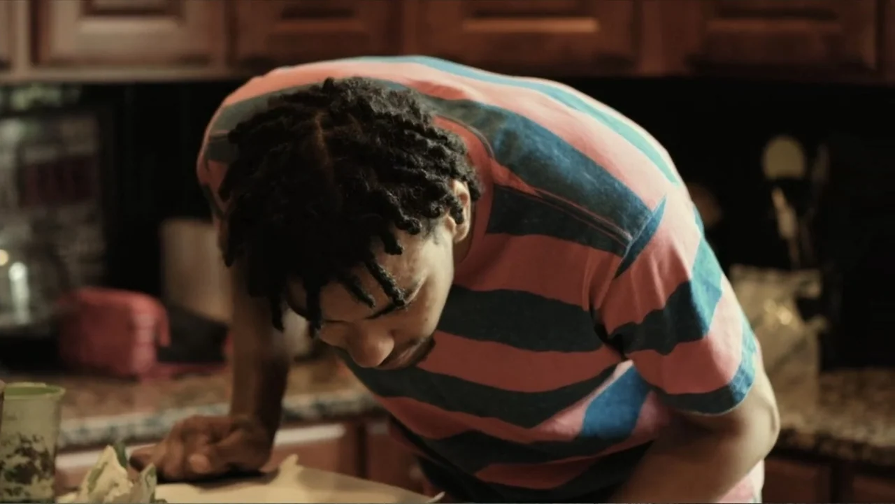 Death of a Child (2017) movie still: medium — A dark residential room with blurred kitchen cabinets and surfaces visible in the background. A young…