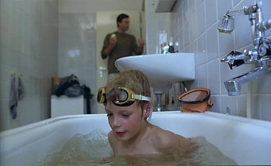 Possession (1981) movie still: medium — A white-tiled bathroom features a child sitting in a bathtub filled with water. A man stands in the…