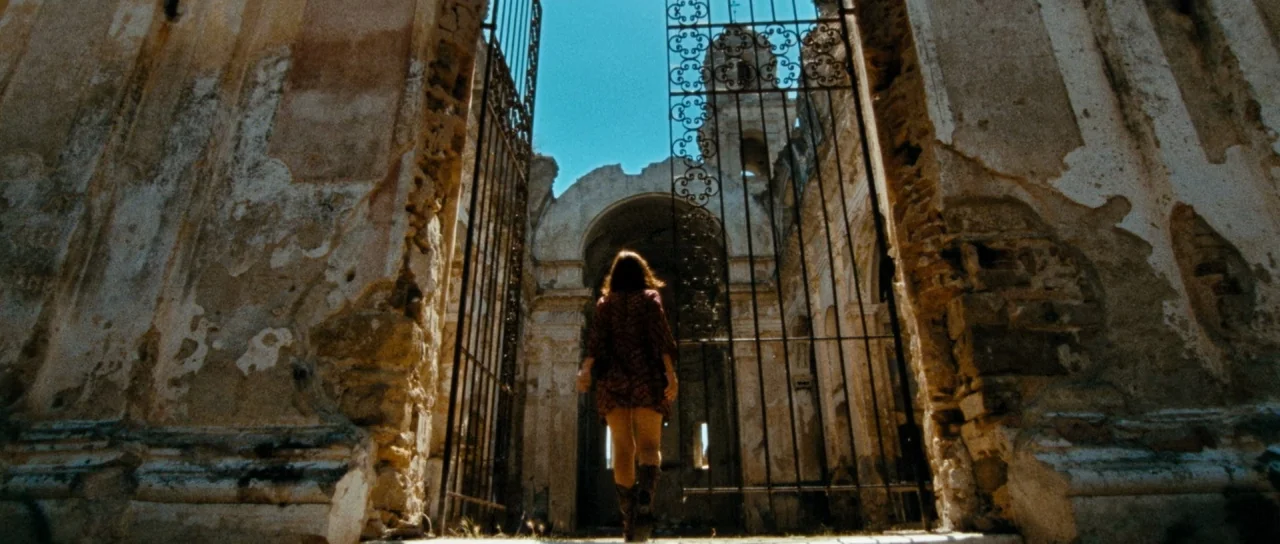 Let The Corpses Tan (2017) movie still: fullbody — An abandoned stone church with collapsed ceilings and crumbling walls stands under a blue sky. A…