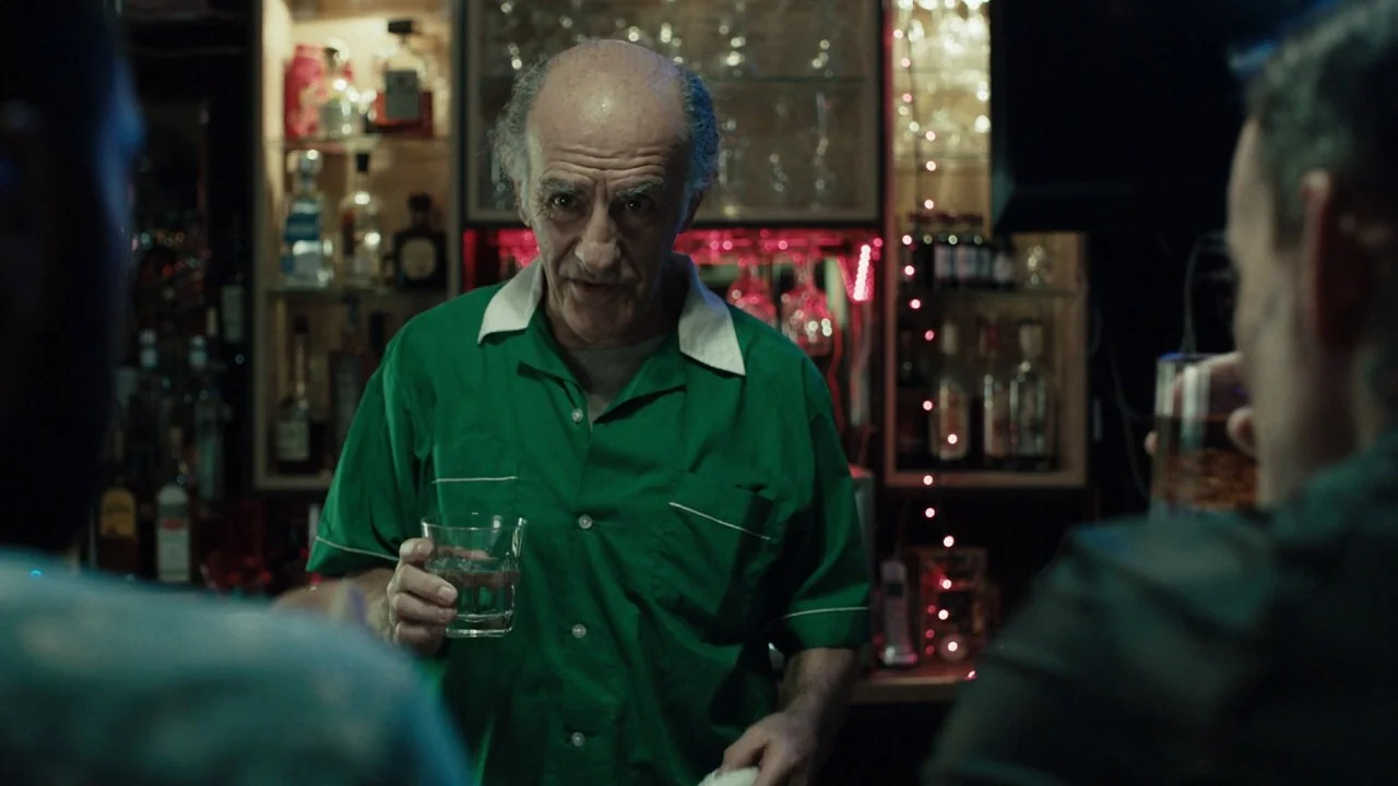 Girl on the Third Floor (2019) movie still: medium — A dimly lit bar setting with backlit shelves displaying various glass liquor bottles. An…