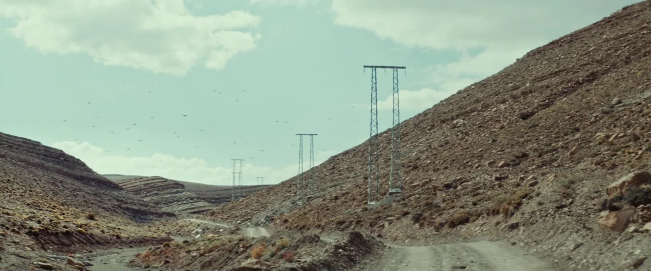 Animalia (2023) movie still: wide — A rugged desert landscape features rocky hills flanking a dry, unpaved dirt road. Metal electricity towers line…