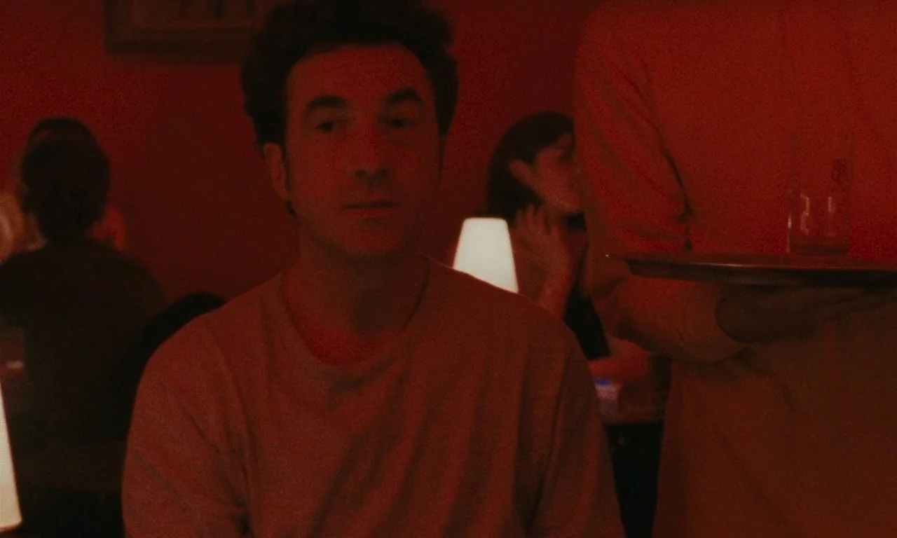 Late August, Early September (1998) movie still: medium — A dimly lit dining space is washed in deep red light from practical lamps. A man sits at a…