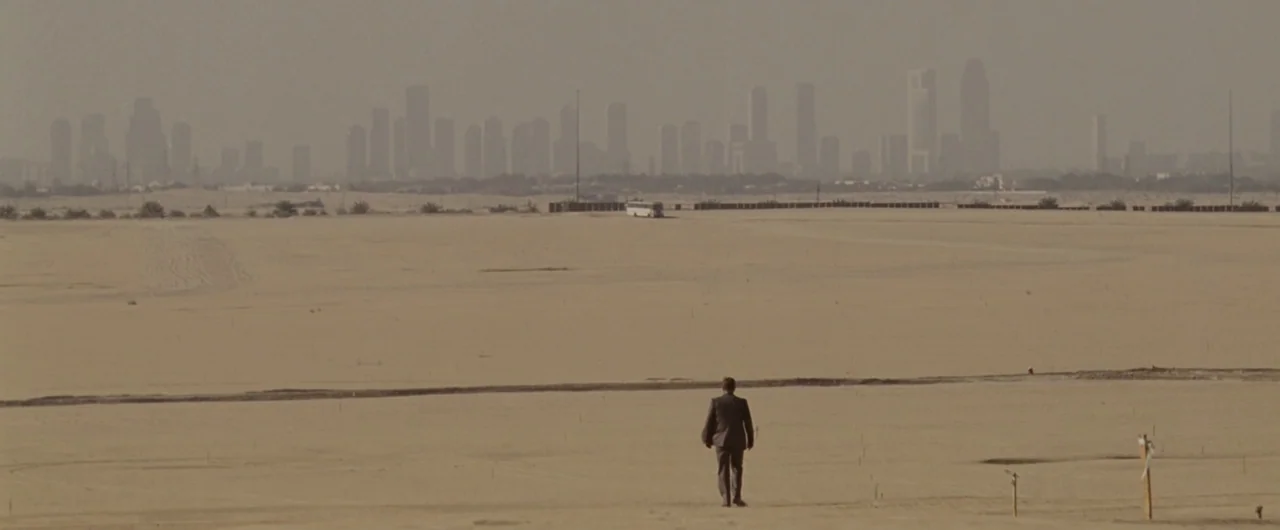 Syriana (2005) movie still: wide — A vast, flat desert terrain stretches toward a distant, hazy modern urban skyline. A lone man in a suit walks…