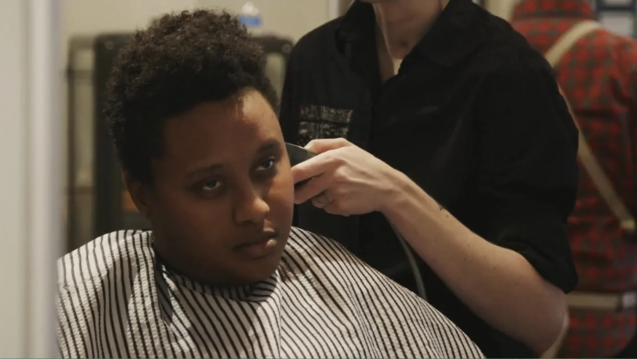 Suited (2016) movie still: medium — A brightly lit interior space with a person getting their hair trimmed by another. The individual in the barber…