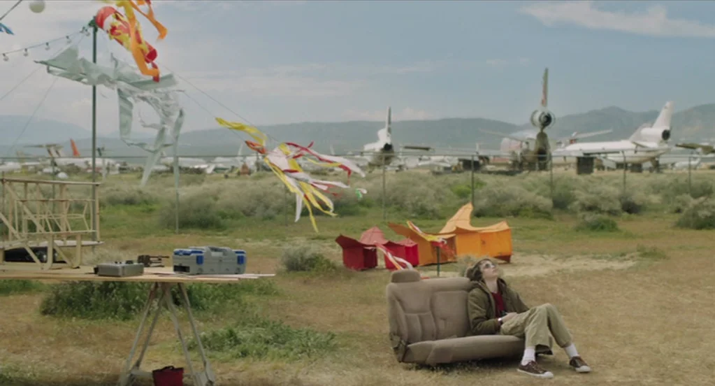 The Congress (2013) movie still: wide — An expansive outdoor airplane graveyard features several grounded aircraft behind a wire mesh fence. A lone…