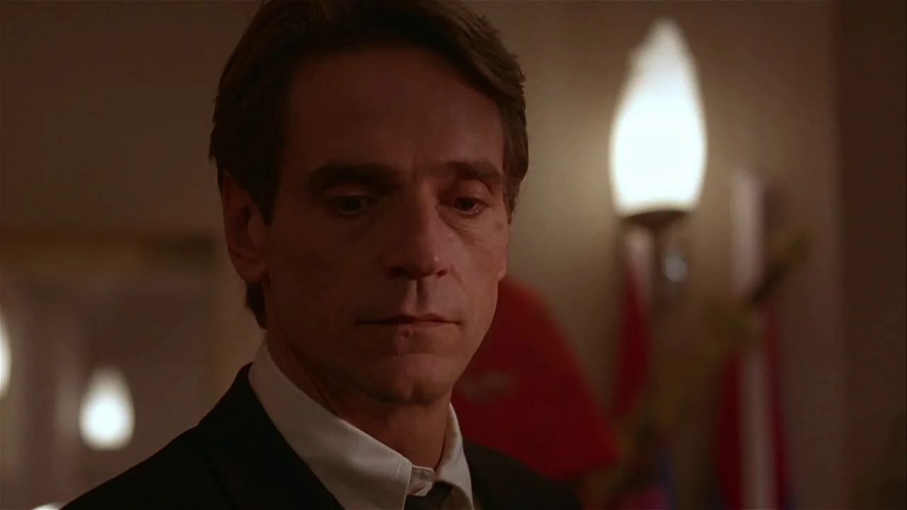 M. Butterfly (1993) movie still: closeup — A dark, dimly lit room contains a wall-mounted light fixture near the subject. The subject in a suit…