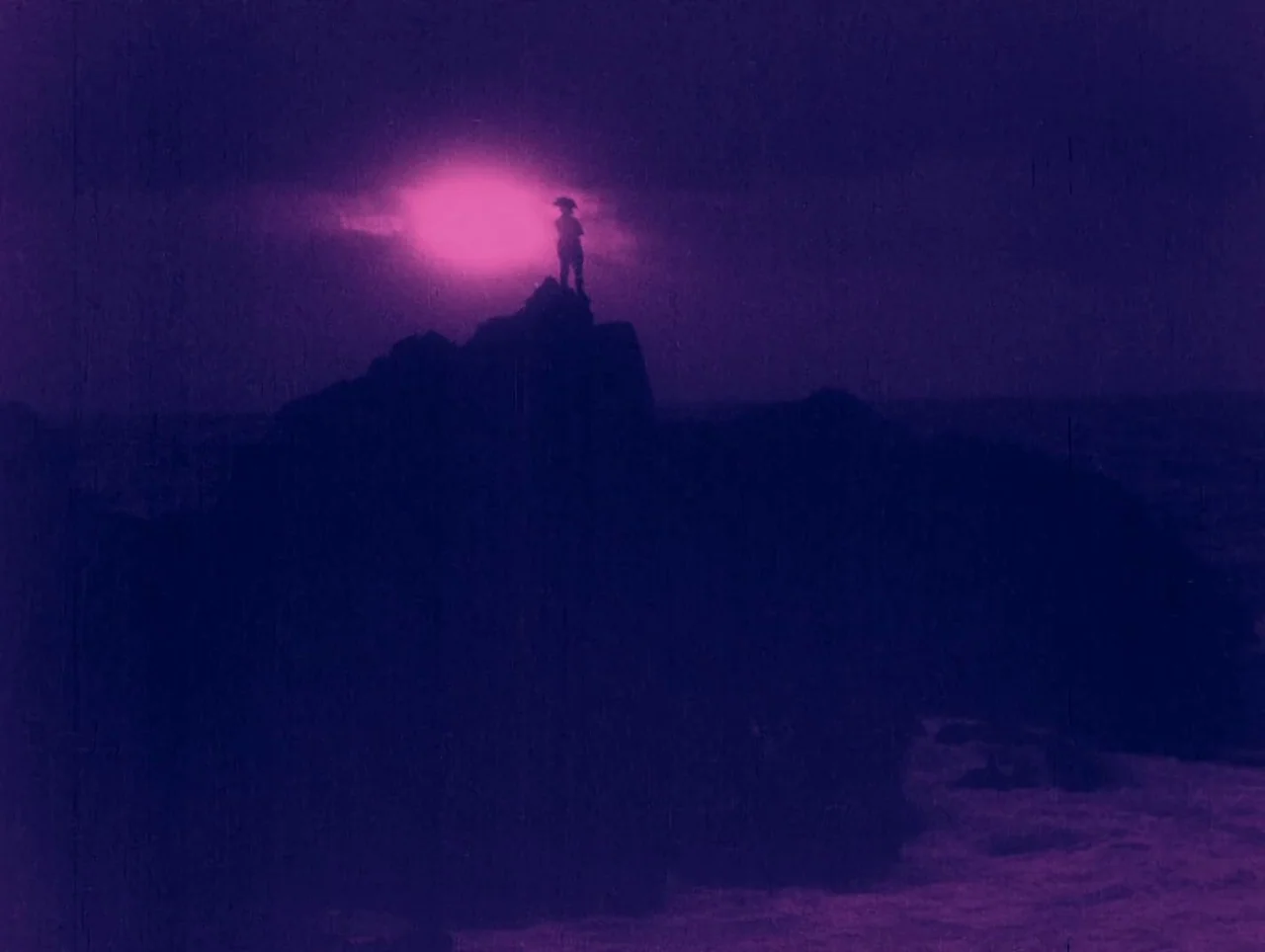 Napoleon (1927) movie still: wide — A dark rocky outcrop rises sharply against a night sky with a glowing orb. A solitary figure stands at the…