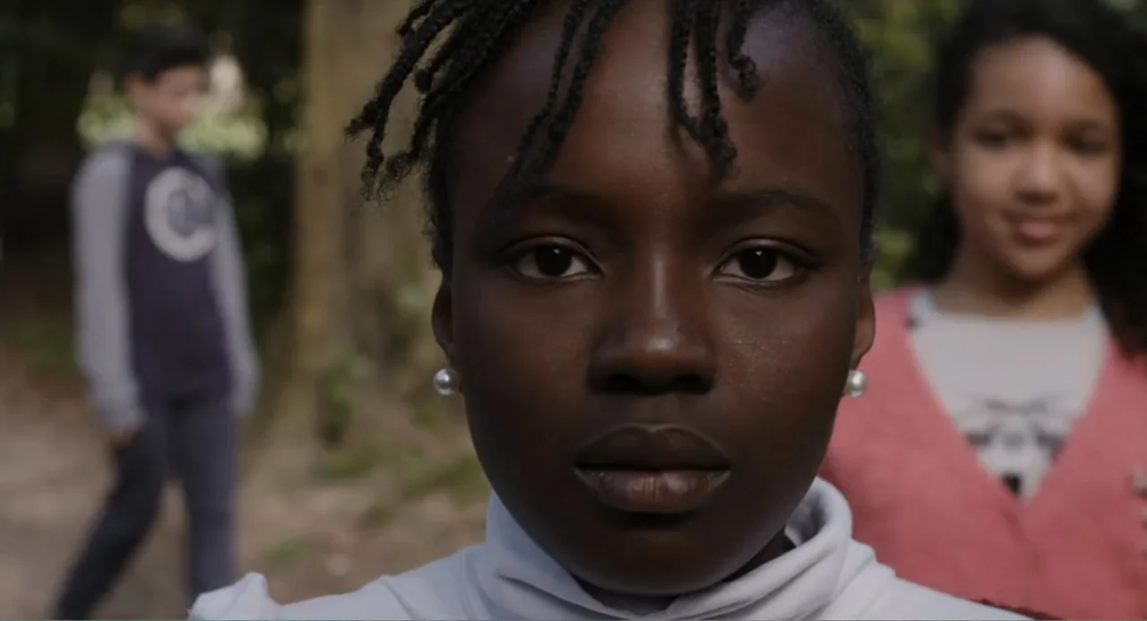 A Dramatic Film (2019) movie still: closeup — A forest trail provides an outdoor setting with blurred foliage visible behind the subjects. A young…