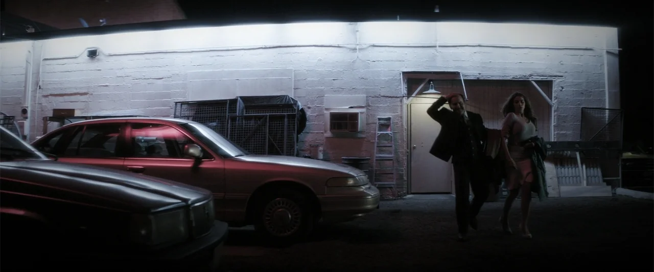Wild Card (2015) movie still: wide — A dark, paved parking area sits before a pale concrete block wall at night. A man and woman traverse the space…