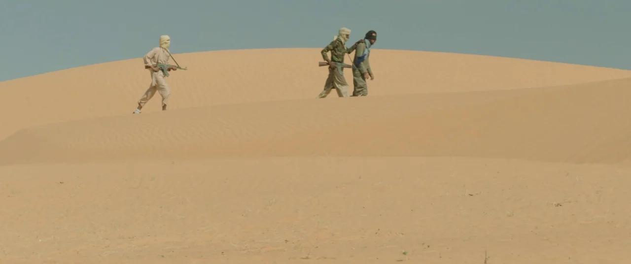Timbuktu (2014) movie still: wide — A vast expanse of rolling golden sand dunes under a pale blue sky. Three men traverse the sandy landscape with…